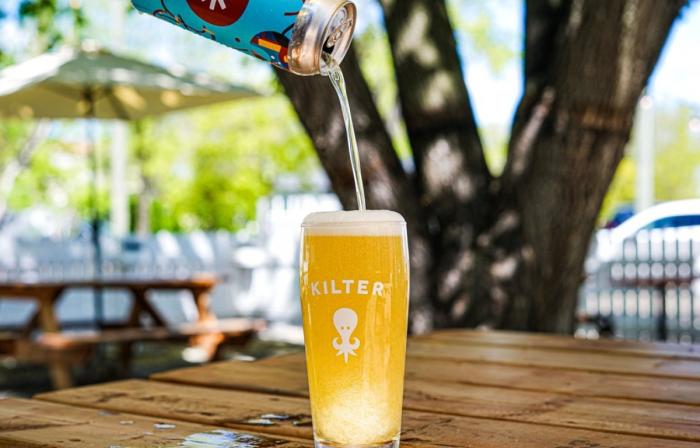 A beer glass on a picnic table in the summer with beer being poured in, the Kilter logo of a squid on front of the glass